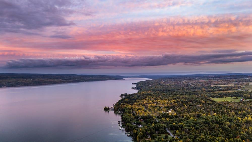 Aerial view down Seneca Lake at sunset with pink clouds, wooded shoreline, and vineyard fields.